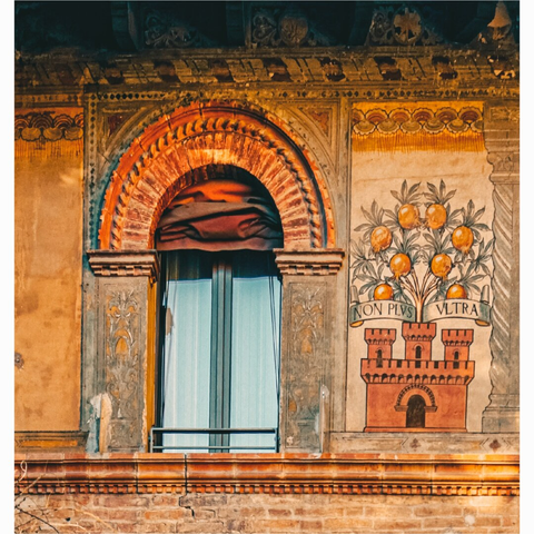 A photo of a window with an arched top made of orange-red bricks, complemented by a header of similar material. The stone pillars on either side of the window bear intricate carvings that have been worn over time. To the right of the window is an interesting detail: a painted emblem featuring a tree laden with golden orange fruits, above a representation of a castle. A banner with the Latin phrase "NON PLVS VLTRA" (Nothing Further Beyond)  is draped across the emblem. The paint on the wall is aged and faded.