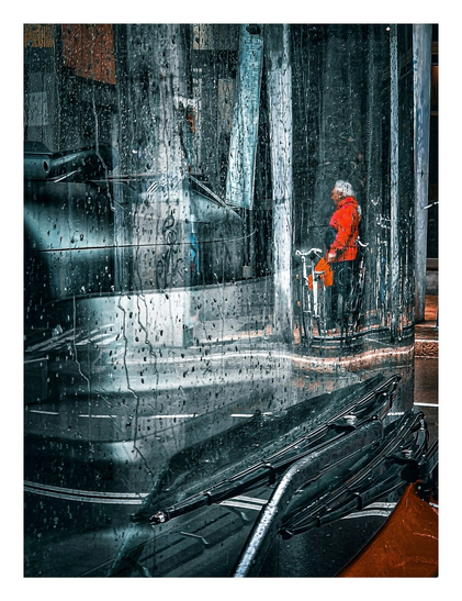 A photo of a rainy street scene viewed through a window reflection, (the windshield of a bus), heavily covered with raindrops. Focus is given to the foreground where numerous raindrops cling to the glass, distorting and blurring the view beyond.
In the lower portion are the windshield wipers lying on the bus windshield. Reflected in the rain-covered glass there is a person standing on the or street close to a row of bikes. They wearing a bright red coat, which stands out against the muted background. They are also holding an orange/red closed umbrella. The background is blurred, with indistinct shapes.