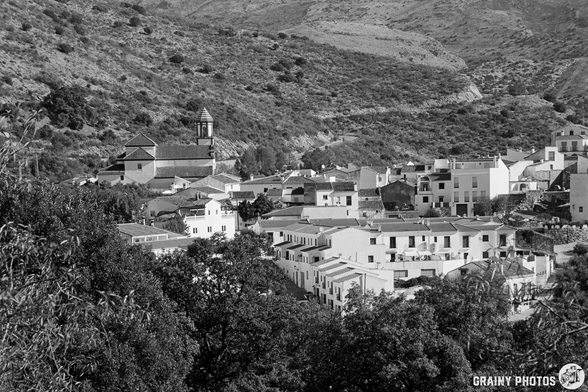 A black and white landscape featuring Atajate nestled in a mountainous region, with traditional architecture, a church tower, dense greenery, and a blend of residential buildings amidst rolling hills.