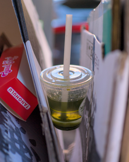 An abandoned plastic drink cup is abandoned in a trash can, wedged in between two sheets of cardboard. The shape of the cup mimics the wedge between the cardboard sides, and the green drink is complimentary to a red food container.