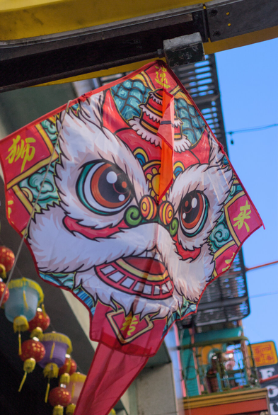 A close up of a red kite with a happy dragon (?) face on it, hanging under the sign for the Chinatown Kites store.