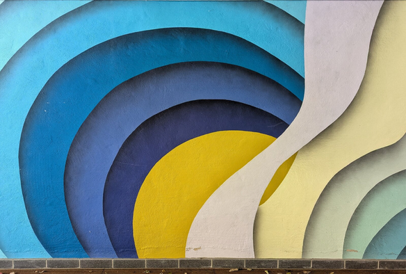 Abstract mural featuring fluid, overlapping arcs in vibrant shades of blue, yellow, grey, and teal. The design creates a dynamic, layered effect with smooth, gradient transitions between colours. The arcs appear to ripple outward from the left, forming a sense of movement and depth, while the bottom edge of the mural aligns with a horizontal brick border. The composition evokes a modern, geometric aesthetic with a focus on bold colour contrasts and organic, flowing shapes.
