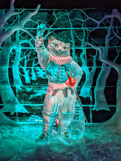 Intricate ice sculpture of Puss in Boots, the fabled feline character, set against a backdrop of icy trees and branches. The sculpture captures the anthropomorphic cat standing upright, dressed in a detailed jacket, breeches, and boots, with one paw raised in a playful or dramatic gesture. A smallsword with a red hilt is strapped to the cat’s belt, adding to its adventurous appearance. The sculpture is illuminated with blue and teal lighting, enhancing the translucent, crystalline quality of the ice. The background features a forest-like design, also carved from ice, creating a whimsical and enchanting scene.