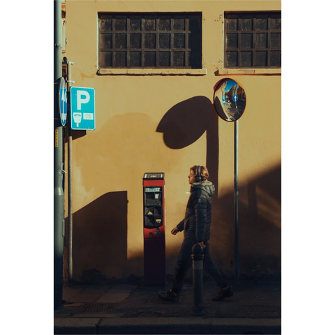 A photograph of a street scene in warm sunlight. A person with brown/blonde hair is wearing a dark blue-grey puffer jacket and dark pants. They are walking past a red parking meter against a yellow, textured wall. There is a blue "P" parking sign (on the right) and a round traffic mirror on the wall (on the right). Shadows are cast, creating a contrast between light and dark areas. The wall has small, grid-like windows near the top of the photo.