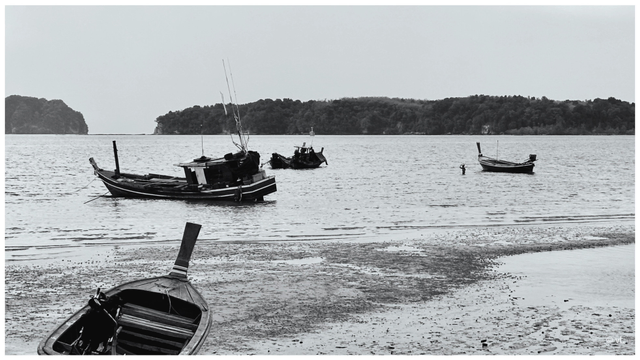 Une port à marrée basse, des barques thaïlandaises échouées sur le sable, au loin une personne dans l’eau jusqu’à la taille porte un paquet sur la tête. En arrière plan, des montagnes. 