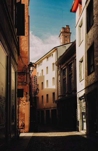 A photograph of a narrow, sunlit Bolognese alleyway/street (Via de' Monari). The alley is flanked by aged, multi-story buildings with textured facades, one in white and another in earthy tones. A black lantern hangs from a orange building on the left, casting shadows. Graffiti is visible on the left building's window. The cobblestone street leads to a central, taller building with a brick chimney. There is a bright blue sky above.