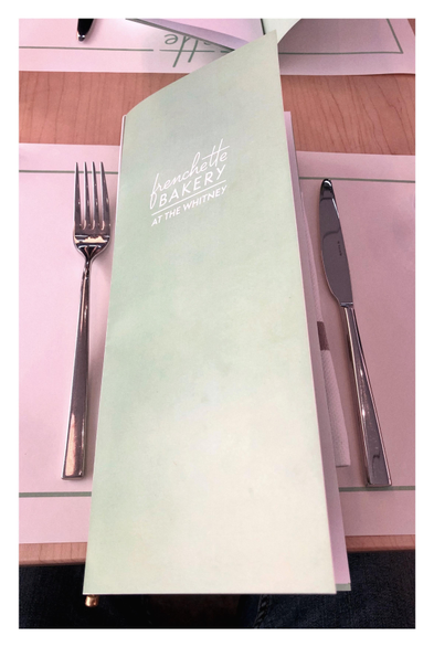 View looking down at a restaurant table (at Frenchette Bakery inside the Whitney Museum in Manhattan). A metal fork and knife, simple but elegant, are placed atop a pink paper placemat with a green border. In between them is a long rectangular menu with a cover slightly lifting upwards. The cover is plain, with a gradient of pale blue-green lightening towards the bottom, and displaying the words “Frenchette Bakery at the Whitney” in stylish font. To me the menu evokes the shape of a skyscraper with a slanted roofline.