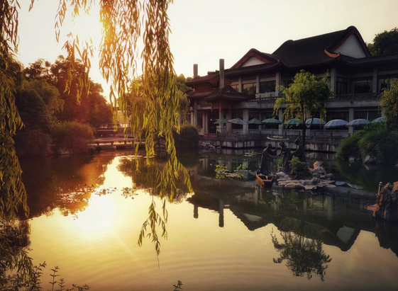 A tranquil sunset scene at a traditional Chinese garden near West Lake, featuring a calm pond reflecting the warm golden light. The foreground is dominated by a serene water body, framed by lush greenery and graceful willow branches that gently cascade into the water. In the background, traditional Chinese architecture with ornate, upturned roofs and wooden structures adds a sense of timeless elegance. The buildings are partially framed by trees and foliage, enhancing the peaceful atmosphere. A stone pathway and small bridges connect the garden areas, while a sculpture of a group of people enjoying a boat ride on the pond adds a touch of artistic charm to the serene landscape.