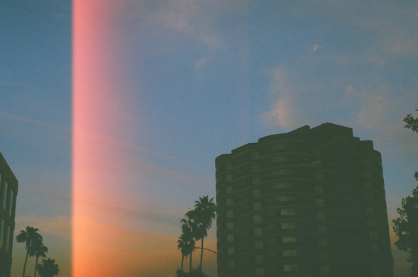 The sunset dips into the skyline over West Hollywood corporate buildings. The film image is distorted with a bright pink light leak.