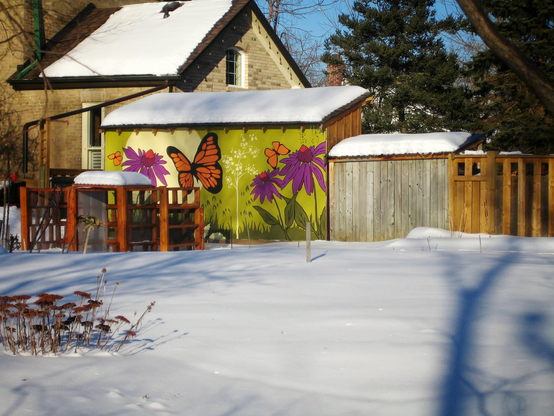 A large butterfly and purple flowers are painted on a green shed connected to a house. Snow covers the ground and large trees can be seen in the background.