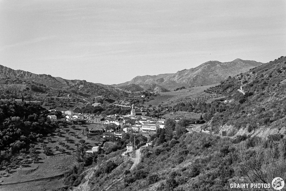 A panoramic view of Atajate nestled in a valley, surrounded by rolling hills and mountains. The scene features a mix of greenery and structures, set against a clear sky in a monochrome style.