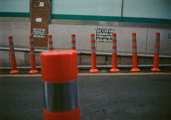 주황색·반사판 볼라드 너머로 도로와 건물이 만나는 땅에 주황색 볼라드가 촘촘하게 박혀있는 것이 보인다. 바로 뒤 건물 벽에는 “잦은 신고로 인해 유튜버/BJ 등 '앉아서 방송' 불가합니다”라는 경고판이 여러 개 붙어있다.
