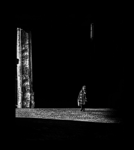 A black and white photograph of a solitary person standing in a dimly lit, shadowy space. The person, wearing a coat, a scarf and dark pants, is centered, casting a long shadow on a textured stone ground. To the left, a tall, vertical column (the front of the church) with a rough, textured surface is illuminated by a narrow light beam. The background is almost entirely black except for a window near the top right.