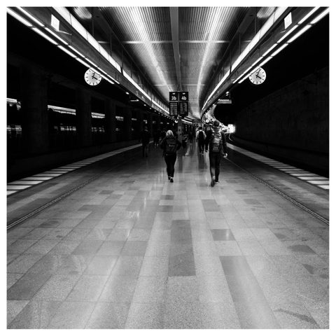 En fyrkantig svartvit bild på en perrong inomhus med två järnvägsspår på sidorna. Två klockor hänger på var sida. Ett tåg på vänster sida och människor går bort över.