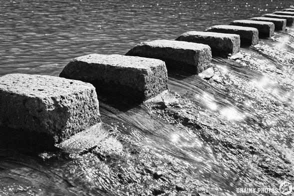 A black and white image of large, rectangular stepping stones arranged in a line across the river Guadiaro, partially submerged in water.  with cascading water and reflections visible on the surface, captured in a monochromatic style.