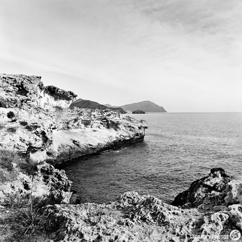 A black and white photo capturing a rugged coastline, featuring rocky cliffs, gentle waves lapping at the shore, and distant mountains under a clear sky. The scene evokes a peaceful yet dramatic natural landscape.