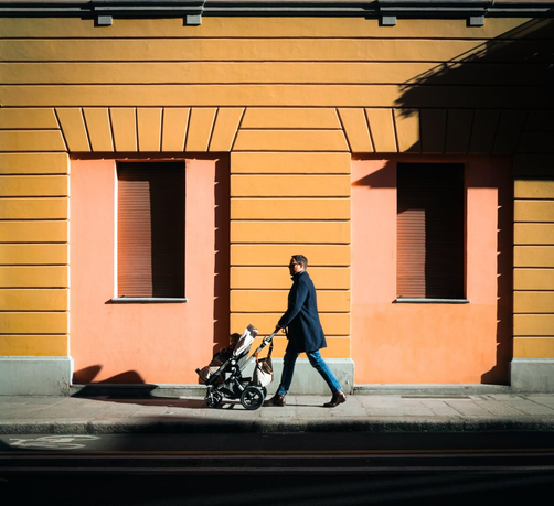 A photograph of a man walking with a buggy on a sunlit pavement in front of an orange-yellow building with two brownish shuttered windows. The man, wearing a dark blue coat, blue jeans, and brown shoes, is pushing a black and white patterned buggy. He has short dark hair and a beard. The building's textured facade casts dark shadows. The street in the foreground has a visible curb.