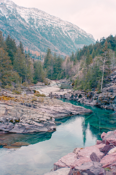 Color photo of a mountain creek zig-zagging its way to the camera carving up ancient sedimentary roks that have a reddish hue. There are many trees framing the roccky banks of the creek and leading the eye towards a big moutain with snow powdered top, barren ridges, and ravines with trees. The sky is a featureless gray which is in stark contrast to the rich emerald waters of the vreek. Shot with a Voightlander BessaR2 and 7Artisan35f2 lens, on KodakEktar100, and developed with KodakC41 by Shom Bandopadhaya. Licensed under Creative Commons Attribution-NonCommercial-ShareAlike (CC BY-NC-SA).