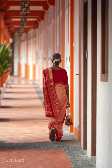This is a colour street photo in portrait format of an Asian lady in a saffron coloured sari (saree) walking along a covered walkway which has the woodwork painted a saffron colour. Chinatown, Singapore (2012).

The camera is looking along a long colonial-era covered walkway that fades out of focus as it bends to the left in the distance, about ten metres away. On the right side is the plastered wall of the front of a restored historic two floor low-rise mixed use: some business, some residential building. Inset into the wall are doors and windows, but with the camera positioned close to the wall, the doors, windows and interiors cannot be seen. The floor is mainly red terracotta tiles with grey stones surrounding the terracotta. On the left are vertical wooden pillars supporting the building overhead the walkway. There is a single green, bushy plant between two of the pillars. The pillars, overhead beams and walls on the right are painted a rich saffron colour. Walking away from the camera and along the right side, close to the wall is an Indian lady wearing an ornate saffron and gold coloured sari, coordinating perfectly with the building she is walking past. 
