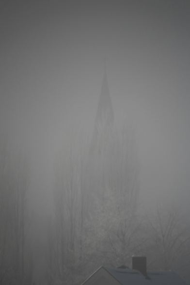 A naturally b&w picture of a church tower behind some trees, engulfed in thick haze. The sun has come over the hills and the light makes the white frost on the trees shine white and the stems stick out.