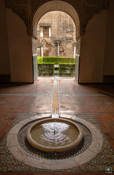 Color photo of a low, circular fountain set in a floor surrounded by decorative tiles. The fountain is inside a room and has a straight outlet channel leading away from it and out through an arched doorway opening leading to a courtyard beyond. 