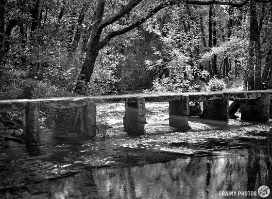 A black-and-white film photo of a simple stone footbridge across a small river in a nice forest setting