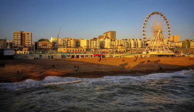 A stunning sunset view of Brighton beach, captured from the vantage point of Brighton Pier. The golden light of the setting sun illuminates the pebble beach, where a few people are scattered, enjoying the serene evening atmosphere. To the right, a large Ferris wheel stands out against the skyline, adding a distinctive touch to the scene. Behind the beach, a row of buildings, including hotels and residential blocks, lines the promenade, their facades bathed in the warm glow of the sunset. The calm sea gently laps against the shore, creating a peaceful and nostalgic ambiance.