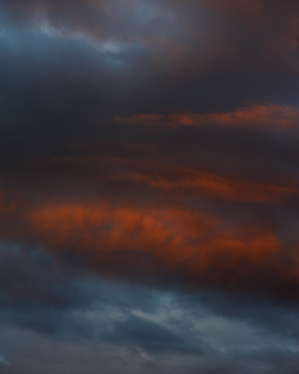 A vertical, dramatic photograph of a cloudy sky around sunset. some clouds are blue-grey, almost with a view of the sky breaking through, and the ones in the middle have an intensely orange colour, almost like glowing.

The clouds are full of contrast and structure, forming large horizontal bands.