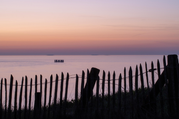 Wir stehen hinter einem einfachen Holzzaun mit vertikalen Latten, die durch Eisendraht zusammengehalten werden, und schauen auf das Meer. Der Zaun hebt sich schwarz vom Meer ab. Die Sonne ist untergegangen und der Himmel ist orange verfärbt. Auch das Meer hat eine orange-lila Farbe. 

We stand behind a simple wooden fence with vertical slats held together by iron wire and look out at the sea. The fence stands out black against the sea. The sun has set and the sky is orange, also the sea has an orange-purple color. 