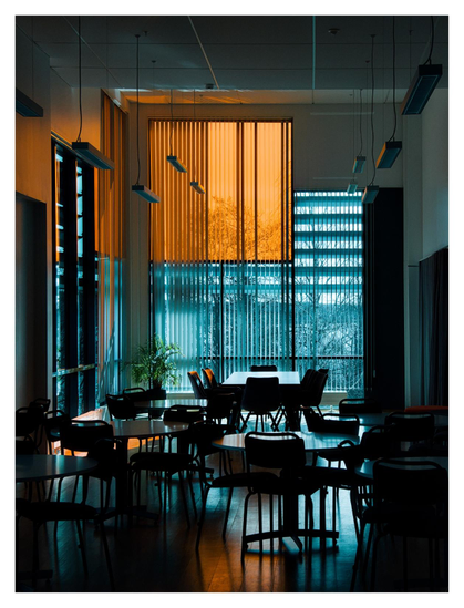 En stående bild på ett rum med 4 bord och många stolar. Längst bort är ett stort fönster i blå ton som delvis täcks av ett fyrkantigt oranget draperi.