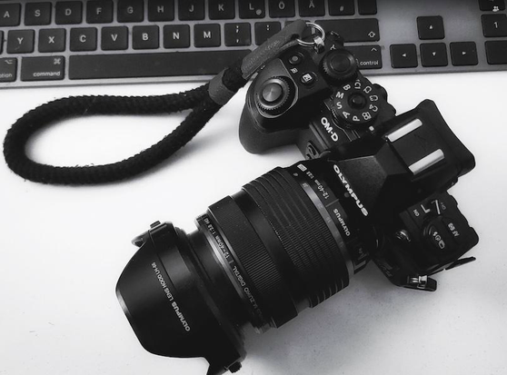 Auf dem Foto sieht man eine Olympus OM-D E-M 1 Mark II mit einem Olympus 12-40mm F2.8 Objektiv. Am oberen Bildrand sieht man einem Computertastatur. Das Foto ist in Schwarzweiß.
