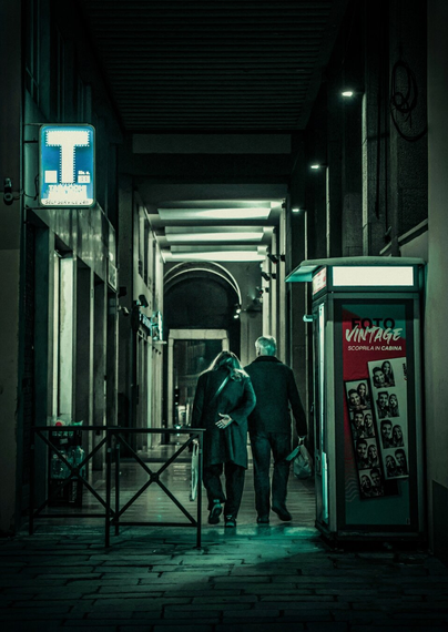 This is a dimly lit, narrow street. There is a couple walking away with their backs to us pretty much in the center of the frame. They are beginning to walk into porticos - which are repeating archways.
To the left, a railing separates the walkway from a darker area. On the righ of the walkway there is a photo booth with images displayed. A blue "Tabacchi" sign is faintly illuminated near the entrance of the walkway ón the left.