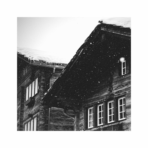 a square black and white image showing a detail of some old wooden houses and their roofs. the dark wood builds up a strong contrast to the white window frames and the fresh snow that's falling