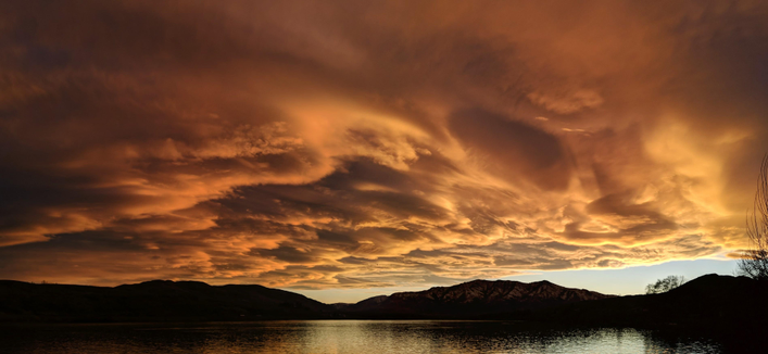 The western sky at sunset in north central washington state usa on February 25, 2026. The clouds appear orangish or reddish and there is clear sky to the right of an elongated mountain peak centered on the right third of the photo. There is slightly rippled water in the foreground.
