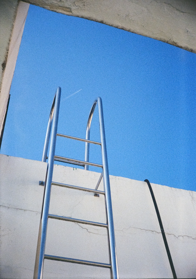 콘크리트 구조물 위로 올라가는 철제 사다리 뒤로 새파란 하늘이 보인다. 사다리 손잡이 사이로는 저 멀리 지나가는 비행기 구름이 작게 보인다.