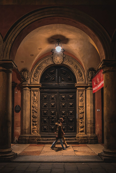 The photo shows a two people walking in front of an ornate, arched entranceway at night. (L'Archiginnasio) The photo has a large, dark wooden door with raised square patterns which are flanked by intricately carved stone pillars and archways. There is warm light from a lamp above the entrance casts shadows and highlights the details of the architecture. A red banner hangs on one side, (signage for the library).
