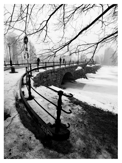 En stående bild på en gammal stenbro med några människor som promenerar på den. Älven är isbelagd med snö och husen i bakgrunden försvinner lite in i diset.