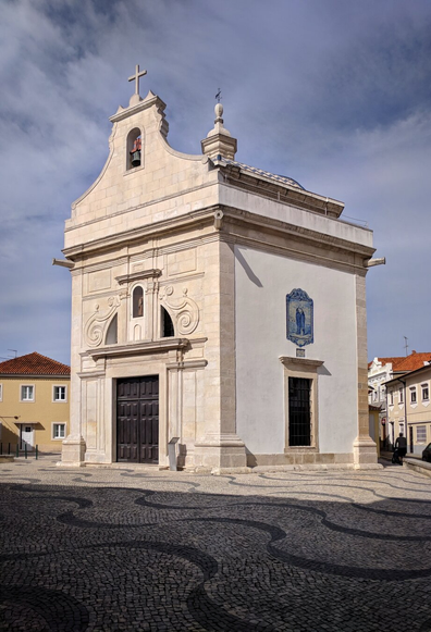 This image depicts the Capela de São Gonçalinho, a charming chapel located in Aveiro, Portugal. The chapel features a compact, square structure with a clean, white façade accented by light-coloured stonework around the windows, door, and corners. The entrance is framed by two sturdy columns, supporting a decorative pediment above the wooden double doors.
A small bell tower, topped with a cross, rises above the chapel’s roof, adding to its quaint and traditional appearance. On the right side of the façade, a blue and white azulejo tile panel depicts a figure, adding a touch of Portuguese artistic heritage. The chapel is set against a backdrop of clear skies and is surrounded by cobblestone pavement with intricate wave-like patterns, enhancing the historic and picturesque ambiance of the scene.