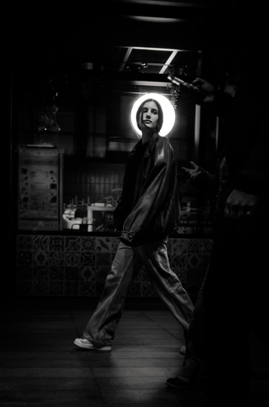The image is a black and white, low-key photograph with dramatic lighting. The primary subject is a young woman walking towards the viewer, positioned slightly left of center. She has dark, shoulder-length hair and is looking directly at the camera with a neutral expression. She is wearing a dark leather jacket over a lighter-colored shirt and loose-fitting pants. Her footwear appears to be sneakers.

Behind her, a bright, circular light source creates a halo effect around her head, making her stand out from the dark background. This light seems to emanate from a window or display behind her. The background is dimly lit and appears to be some sort of establishment, possibly a cafe or shop, with signs and tiled walls visible. The tiles have a repeating pattern.