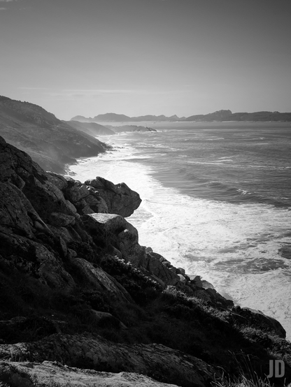La imagen muestra una costa rocosa con acantilados que descienden hasta el mar. Las olas rompen contra la orilla, creando espuma blanca que contrasta con el agua oscura. Al fondo se ven montañas y una línea de horizonte clara bajo un cielo despejado. La foto está en blanco y negro, lo que resalta las texturas de las rocas y el movimiento del mar.