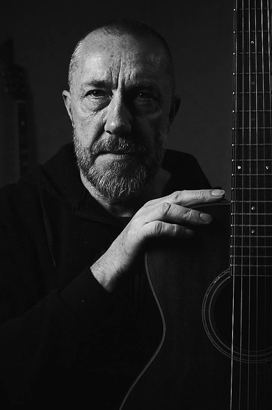This high-contrast, black-and-white, vertical portrait features a man with a weathered, expressive face and a salt-and-pepper beard. He is looking directly into the lens with a soulful gaze. He is wearing a dark hooded sweatshirt which blends into the shadows, drawing the viewer's attention to his face and right hand. His hand rests firmly on the body of an acoustic guitar, held vertically, which occupies the right third of the frame, revealing the intricate details of the strings and fretboard. Dramatic side lighting creates a sharp chiaroscuro effect, highlighting the textures of his skin and the silver in his beard while casting the other side of his face into deep shadow. Against the dark, minimalist backdrop, the faint, blurred silhouette of a guitar neck hints at a dedicated musical space or studio. The overall mood is contemplative and artistic, capturing the intimate and serious connection between a mature musician and his instrument.