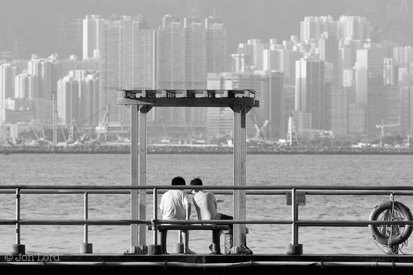 This is a black and white street photo in landscape format of two people sitting on a bench under a small storm shelter overlooking the water. North Point, Hong Kong (2014).

Stretching across the frame from left to right are two sets of parallel metal railings on a small concrete pier with a life ring attached on the far right side. In the centre is a small bench, no more that three people wide that is under an equally small storm shelter. Sitting on the bench are two young men, facing away from the camera and leaning towards each other as if looking at something that's between them. Beyond them is a body of medium grey harbour water that rises upwards to the halfway up level. At that point there is a stone, harbour breakwater, again running across the photo from left to right. behind the breakwater, though blurred, are the numerous cranes of moored derrick barges. Behind is a scene of a densely spaced cityscape with multiple white high-rise apartment blocks. 

The pier is at North Point, Hong Kong. The water is Victoria Harbour. The breakwater is the To Kwa Wan Typhoon Shelter and the cityscape is Kowloon.