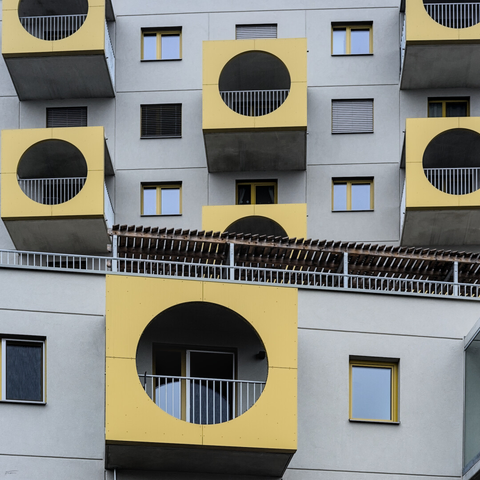 Das Foto zeigt die Fassade eines Wohngebäudes. Auffällig sind die großen, quadratischen Balkonbereiche mit gelben Paneelen und runden Öffnungen. Die Balkone haben weiße Geländer. Im Hintergrund verläuft eine Brücke oder Überdachung. Die Fenster des Gebäudes sind schlicht und in neutralen Farben gehalten. Die Architektur wirkt geometrisch und farbenfroh.
 ---- 
The photo shows the facade of a residential building. The large, square balcony areas with yellow panels and round openings are striking. The balconies have white railings. In the background, there is a bridge or canopy. The windows of the building are simple and neutral in color. The architecture appears geometric and colorful.