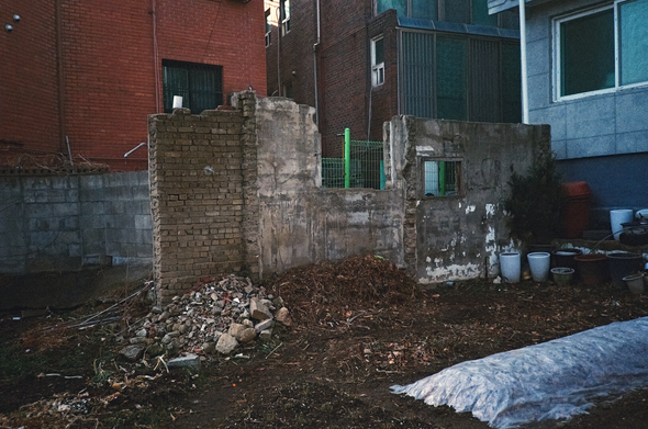 주택가 공터에 폐허 일부가 담장처럼 벽 한 쪽만 남았다. 창문 두 개 중 큰 것은 위쪽이 완전히 뚫린 상태가 되었다.