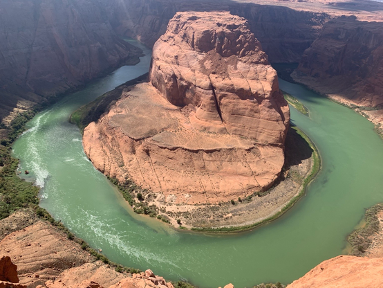 A large, reddish-brown rock formation rises in the center of a winding green river, surrounded by steep canyon walls and arid terrain. The scene captures the natural beauty of a desert landscape.