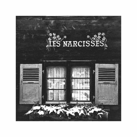 a square black and white image showing an old window with originally blue symmetric shutters in an old wooden house. above the window there's a decorative inscription saying "ies narcisses" together with some paintings of the flowers