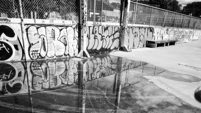 A large pool of water sits on an outdoor basketball court. Graffiti from a surrounding wall reflects in the water in this black and white image.