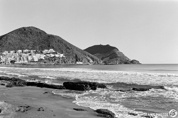 Scenic beach view with gentle waves lapping at the shore, surrounded by rocky formations and hills. A coastal town is visible on the hillside, captured in black and white for a classic and serene atmosphere.