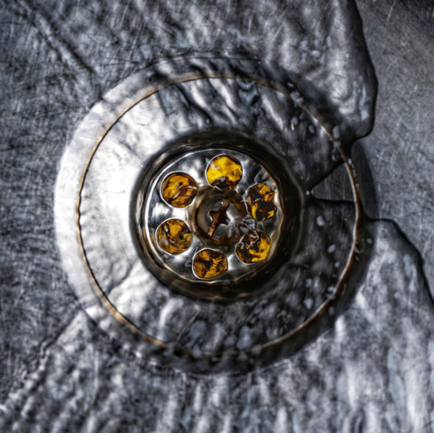 Das Bild zeigt eine Nahaufnahme eines kreisrunden Metallabflusses in einem Spülbecken aus Edelstahl, der von fließendem Wasser umgeben ist. Im Zentrum des Bildes befindet sich das Abflusssieb mit sechs kreisförmig angeordneten Öffnungen, durch die man bernsteinfarbenes Licht  schimmern sieht, was einen starken farblichen Kontrast zum ansonsten silbergrauen Metall bildet. Eine zentrale Kreuzschlitzschraube hält das Sieb in der Mitte fixiert. Das umgebende Edelstahlbecken weist eine stark gebürstete oder zerkratzte Textur auf, über die das Wasser in unregelmäßigen, dynamischen Wellen und Schlieren abfließt. Die Beleuchtung erzeugt auf der nassen Metalloberfläche und den Wasserbewegungen ein komplexes Spiel aus hellen Glanzlichtern und tiefen Schatten, wodurch die Szene sehr plastisch und fast abstrakt wirkt.