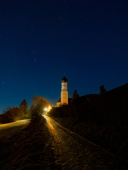This image captures a serene night scene featuring a prominent church under a vast, star-filled dark blue sky. The sky, which takes up roughly the top two-thirds of the image, is densely populated with countless tiny white stars, creating a luminous celestial tapestry. Below the starry sky, a light-colored church with a distinctive domed tower stands out, illuminated by artificial light. A narrow, unpaved path, also lit by a warm, golden light source (likely a street lamp just out of frame to the left), curves upwards from the foreground towards the church. On either side of the path, the ground is covered in dark grass and some sparse trees and bushes, with some patches of what appears to be snow or frost, reflecting the warm light. To the right of the church, a dark, sloped hill covered in vegetation rises, mostly silhouetted against the night sky.